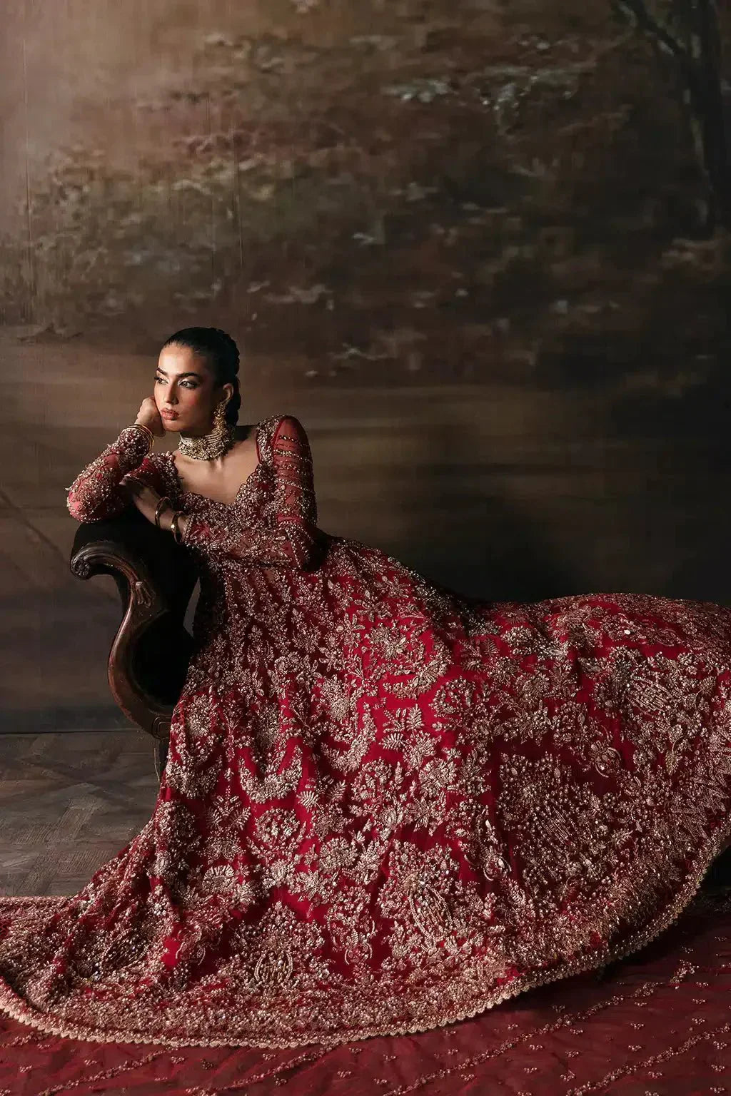 Afrozeh The Brides Edit 23 Adelaide bridal gown in brick red net with gold embellishments, model seated elegantly on a vintage chair.